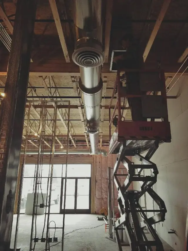 Spiral supply duct and round diffusers hung in an open-joist restaurant ceiling beside a scissor lift